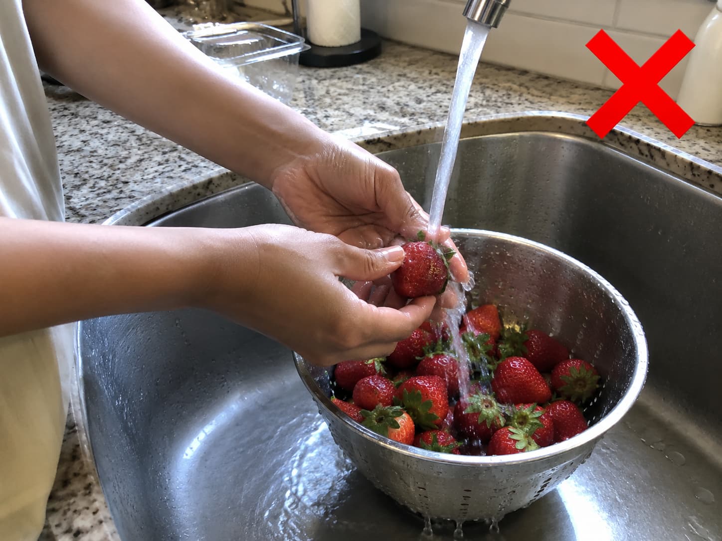 Hands rinsing strawberries under running water with a red X overlay to show they should not be washed before storage